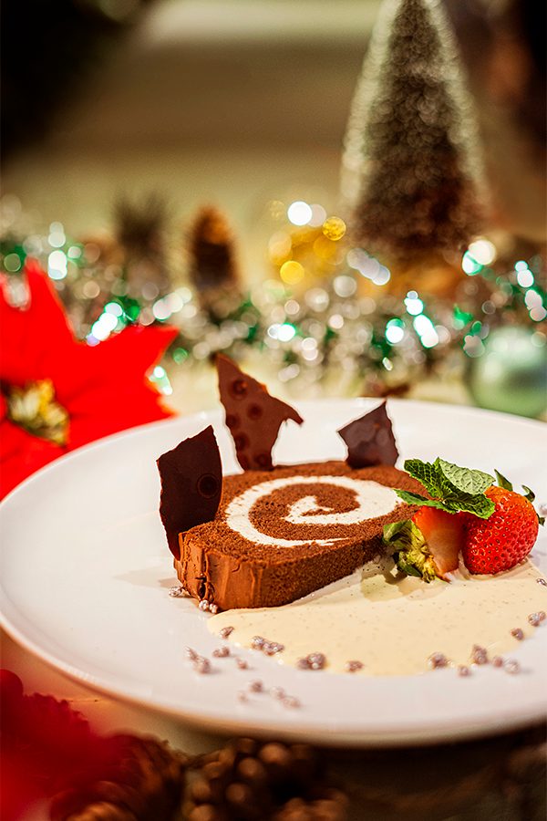 A chocolate vanilla log served with English vanilla cream and strawberries. Christmas decorations are placed around the plate. 